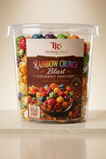 A clear plastic tub of TRS The Royal Treats Rainbow Crunch Blast Gourmet Popcorn filled with multicolored cereal pieces, set on a light beige surface against a warm beige background.