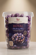 A clear plastic tub of TRS Royal Blueberry Bliss Gourmet Popcorners, filled with purple popcorn and blueberries, set on a light gray marble surface against a warm beige background.